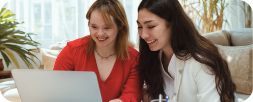 Two young women, one with Down syndrome, smiling while looking at a laptop together — the community your donation supports